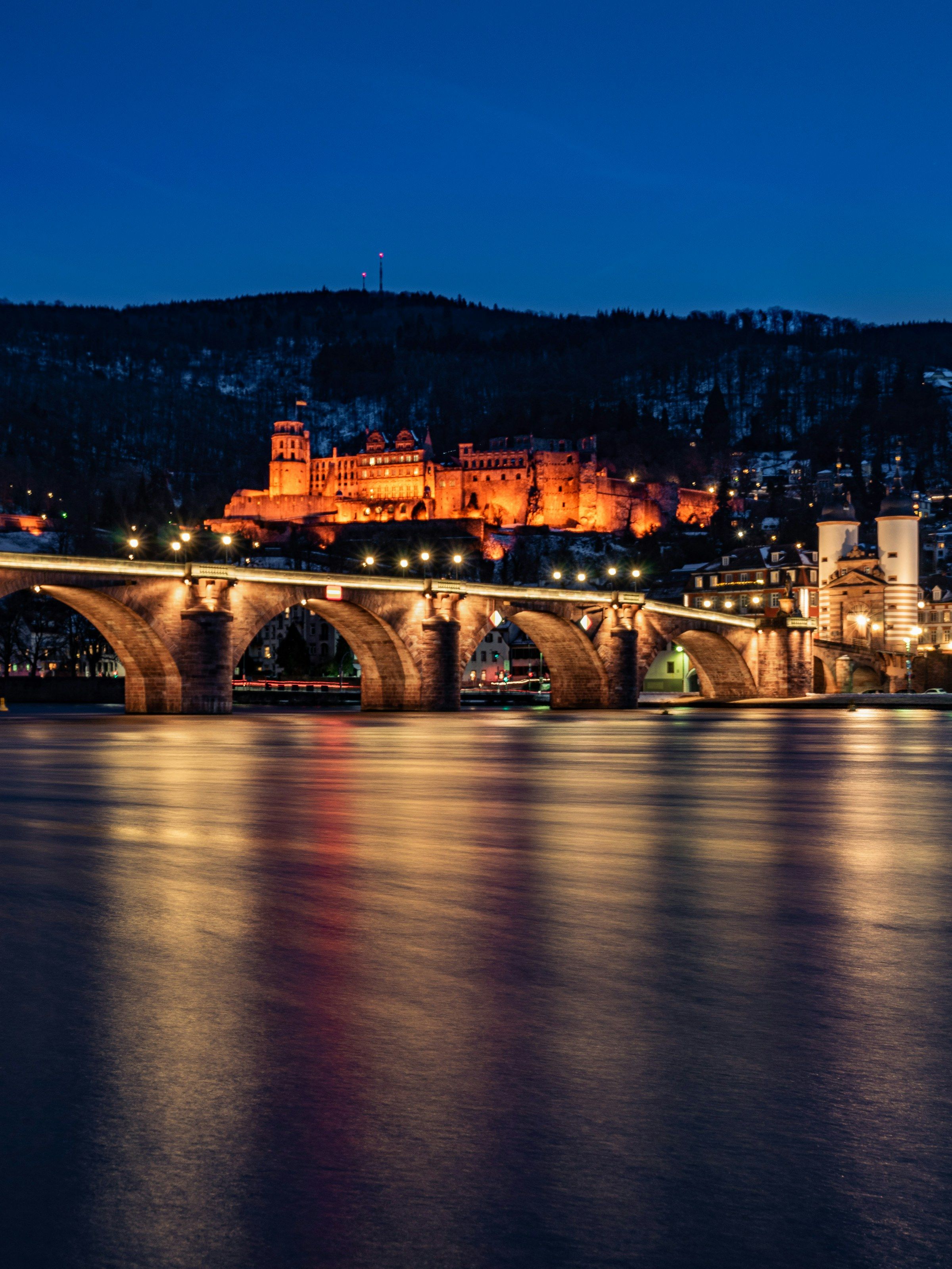Die beleuchtete Alte Brücke in Heidelberg bei Nacht, dahinter das angestrahlte Schloss; die Lichter spiegeln sich im Neckar