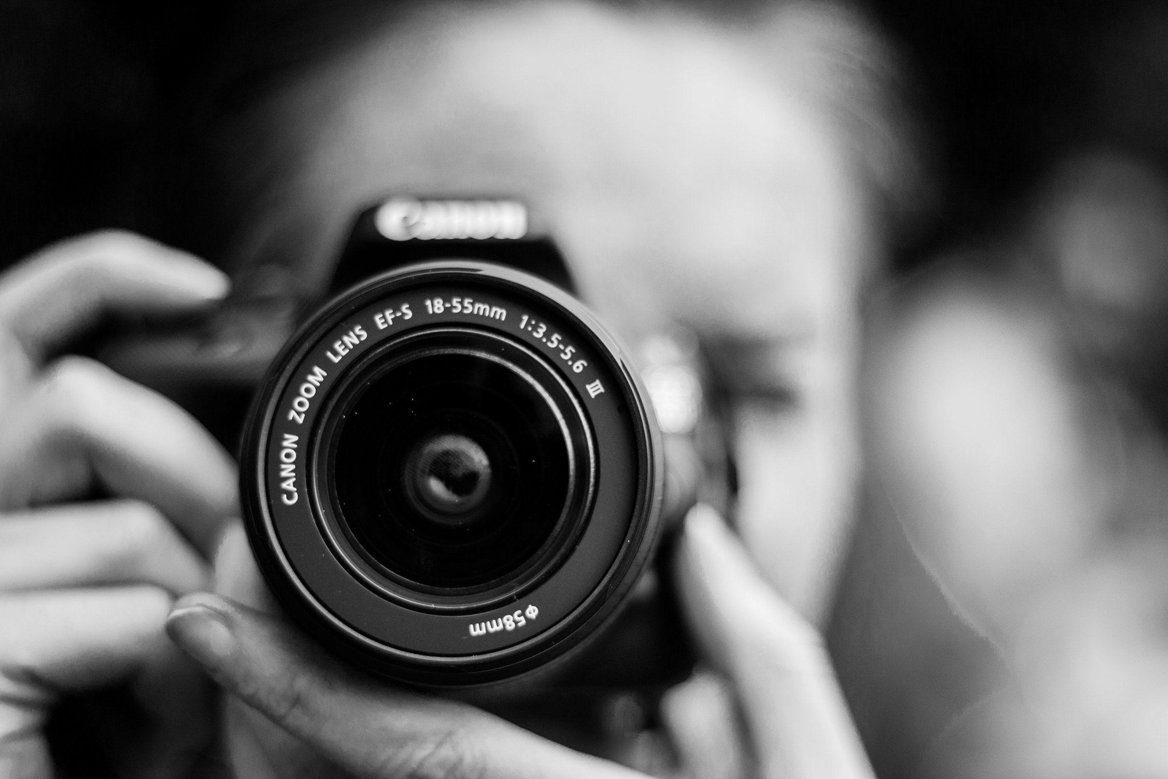 Schwarzweißfoto: Eine Person hält eine Kamera mit Zoomobjektiv direkt in Richtung Betrachter