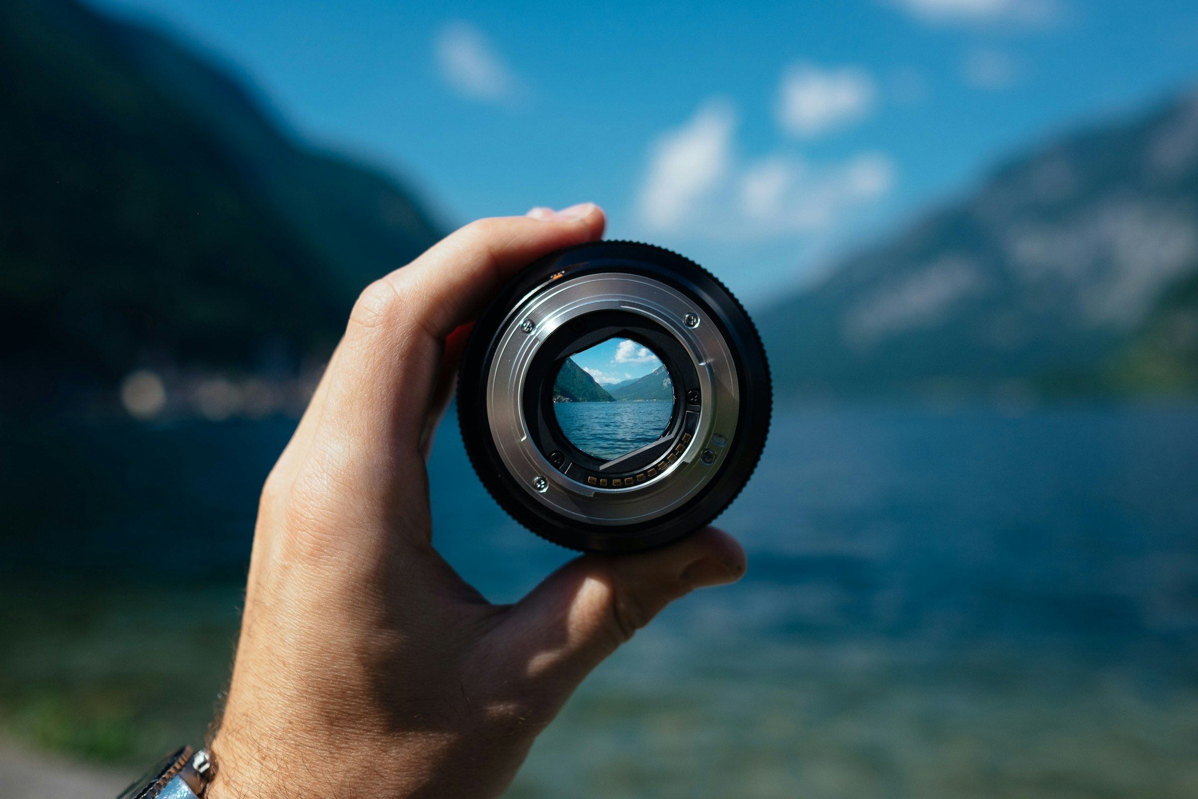 Eine Hand hält ein abgenommenes Objektiv vor einen See, durch das Glas erscheint die Landschaft scharf